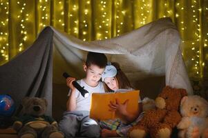 leyendo y familia juegos en para niños carpa. chico y niña con libro y Linterna antes de yendo a cama foto