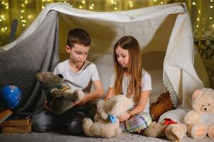 Siblings sit in a hut of chairs and blankets. Brother and sister reading book with flashlight at home photo