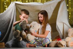 dos pequeño niños jugar a hogar en el noche a construir un cámping tienda a leer libros con un Linterna y dormir adentro. foto