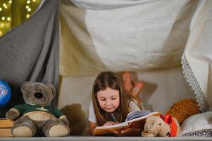 child girl reading with book and flashlight and teddy bear in tent. before going to bed photo