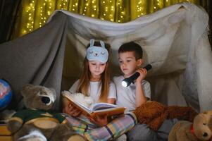 Siblings sit in a hut of chairs and blankets. Brother and sister reading book with flashlight at home photo