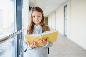 frente ver de pequeño hermosa colegio niña entre corredor a escuela, participación notas a manos. gracioso y contento niña sonriente a cámara, descansando después lecciones en primario colegio foto