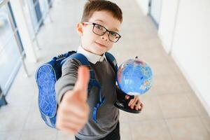 Happy smiling boy in glasses is going to school for the first time. Child with school bag and book in his hand. Kid indoors of the class room . Back to school photo