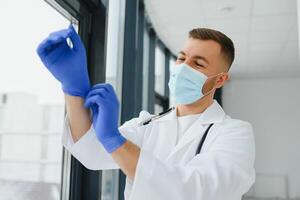 joven sonriente caucásico masculino médico en blanco uniforme, con los anteojos y estetoscopio alrededor cuello poniendo caucho guantes antes de paciente examen foto