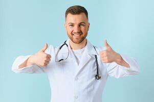 Waist up portrait view of the smiling happy doctor showing big finger at the free space on blue background. Doctor and medicine concept photo