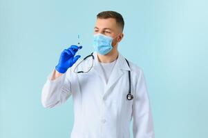 Vaccination against coronavirus. Testing COVID-19 vaccines. A male doctor in a medical mask fills a syringe the with a COVID-19 vaccine on a blue background. Doctor's hands hold a syringe close-up photo