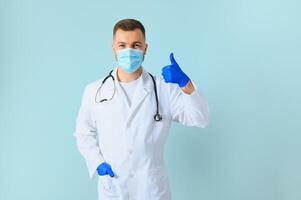 Young male doctor wearing medical mask, and stethoscope round his neck, isolated on blue background photo