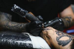 recortado cerca arriba de un barbado tatuaje artista trabajando a su estudio tatuajes manga en el brazo de su masculino cliente. hombre consiguiendo tatuado por profesional tatuador foto
