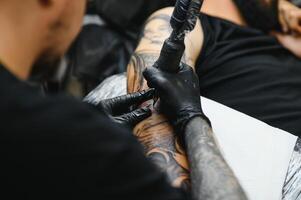 recortado cerca arriba de un barbado tatuaje artista trabajando a su estudio tatuajes manga en el brazo de su masculino cliente. hombre consiguiendo tatuado por profesional tatuador foto