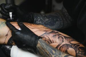 recortado cerca arriba de un barbado tatuaje artista trabajando a su estudio tatuajes manga en el brazo de su masculino cliente. hombre consiguiendo tatuado por profesional tatuador foto