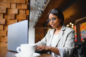 Young African American girl sitting in restaurant and typing on her laptop. Pretty girl working on computer at cafe. photo