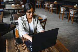 Beautiful african business woman waiting for the meeting in a co-working space and sitting with the portable computer. Hipster girl is surfing the web on a laptop during lunch. photo