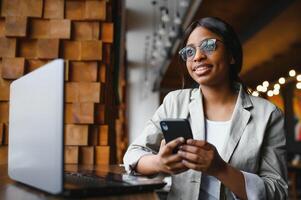 Beautiful african female with a long dark hair is typing messages on a smartphone while sitting beside the window in a soft light. Young business woman is using laptop and mobile phone for the work. photo