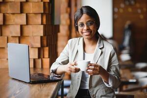 Head shot portrait of happy smiling African American woman sitting at table in cafe, looking at camera, excited female posing, working at computer, doing homework, preparing report in coffee house photo