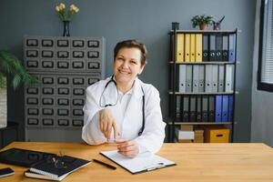 sonriente hembra cuidado de la salud trabajador haciendo algunos papeleo y utilizando ordenador portátil mientras trabajando a del doctor oficina. foto