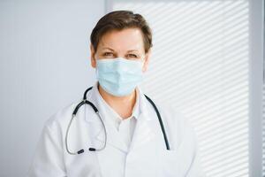Older female doctor wearing face mask and white medical coat standing in hospital. Portrait photo