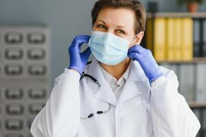 más viejo hembra médico vistiendo cara máscara y blanco médico Saco en pie en hospital. retrato foto