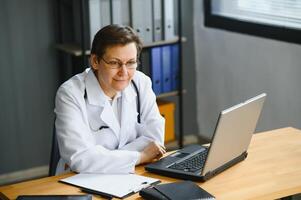 Portrait shot of middle aged female doctor sitting at desk and working in office. photo