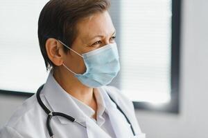 más viejo hembra médico vistiendo cara máscara y blanco médico Saco en pie en hospital. retrato foto