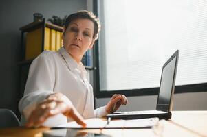 Smiling stylish mature middle aged woman sits at desk with laptop, portrait. older senior businesswoman. photo