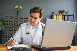 Smiling female healthcare worker doing some paperwork and using laptop while working at doctor's office. photo