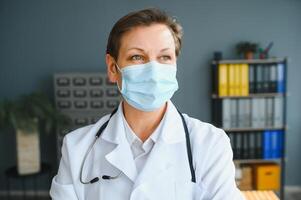 más viejo hembra médico vistiendo cara máscara y blanco médico Saco en pie en hospital. retrato foto