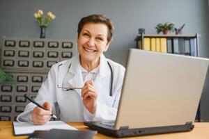 Smiling female healthcare worker doing some paperwork and using laptop while working at doctor's office. photo