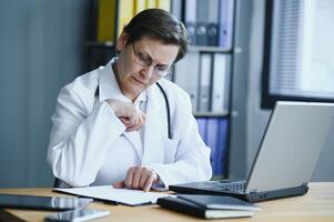 Smiling female healthcare worker doing some paperwork and using laptop while working at doctor's office. photo