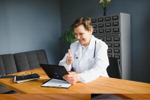 Portrait of female doctor counseling patient via call. Professional physician in white lab coat gesturing and explaining course of treatment sitting at office desk during online consultation. photo