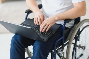 Portrait of smiling disabled male sitting in wheelchair and working on laptop from home. Young worker with special needs. Freelancer and people with disabilities concept photo