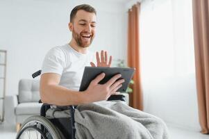 Man in wheelchair using tablet computer at home. Positive retired male with physical disability browsing web on touch pad, watching online indoors. photo