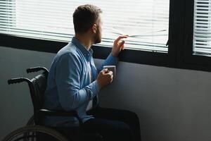 discapacitado persona se sienta en silla de ruedas en contra ventana. grave triste caucásico hombre vistiendo casual ropa y Mira a grande panorámico ver en brillante moderno vivo habitación o hospital. foto