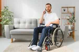 The disabled person sits in a wheelchair. He is talking to someone on his smartphone. He is in his large bright living room. He smiles. photo