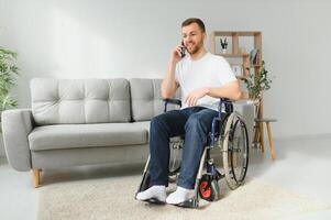 The disabled person sits in a wheelchair. He is talking to someone on his smartphone. He is in his large bright living room. He smiles. photo