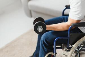 sportsman sitting in wheelchair and outstretching arms with dumbbells during rehabilitation exercise in modern medical center. Man sitting on wheelchair at home photo