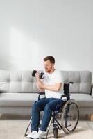 sportsman sitting in wheelchair and outstretching arms with dumbbells during rehabilitation exercise in modern medical center. Man sitting on wheelchair at home photo