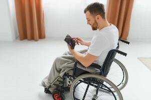 Man in wheelchair using tablet computer at home. Positive retired male with physical disability browsing web on touch pad, watching online indoors. photo