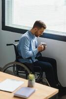 discapacitado persona se sienta en silla de ruedas en contra ventana. grave triste caucásico hombre vistiendo casual ropa y Mira a grande panorámico ver en brillante moderno vivo habitación o hospital. foto