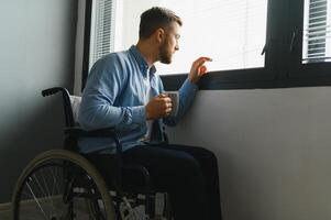 Disabled Person Sits in Wheelchair Against Window. Serious Sad Caucasian Man Wearing Casual Clothes and Look at Large Panoramic View in Bright Modern Living Room or Hospital photo
