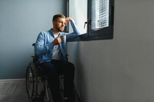 Sad disabled person feeling lonely at home or clinic. Depression on self isolation. Upset man in wheelchair looks out of large window and dreams, in living room interior. photo