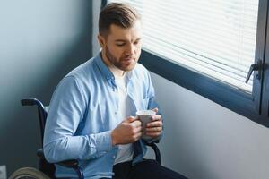 Disabled Person Sits in Wheelchair Against Window. Serious Sad Caucasian Man Wearing Casual Clothes and Look at Large Panoramic View in Bright Modern Living Room or Hospital. photo