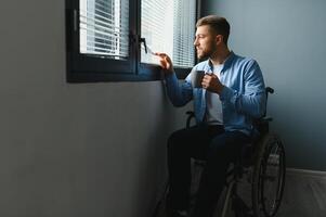 Disabled Person Sits in Wheelchair Against Window. Serious Sad Caucasian Man Wearing Casual Clothes and Look at Large Panoramic View in Bright Modern Living Room or Hospital photo