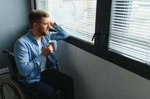 Disabled Person Sits in Wheelchair Against Window. Serious Sad Caucasian Man Wearing Casual Clothes and Look at Large Panoramic View in Bright Modern Living Room or Hospital photo