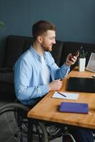 joven contento empresario en silla de ruedas leyendo mensaje en inteligente teléfono mientras trabajando a hogar foto