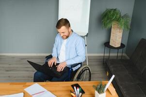 Young man with special needs in casual clothes working on wireless laptop. Male freelancer working from home while sitting in wheelchair photo