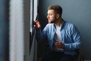 Disabled Person Sits in Wheelchair Against Window. Serious Sad Caucasian Man Wearing Casual Clothes and Look at Large Panoramic View in Bright Modern Living Room or Hospital photo