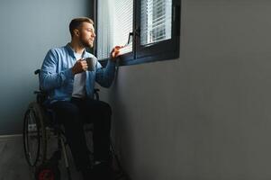 joven discapacitado hombre sentado en un silla de ruedas cerca el ventana. foto