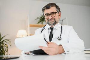 retrato de simpático sonriente mayor adulto cuidado de la salud profesional terapeuta sentado a lugar de trabajo. contento confidente más viejo masculino médico médico vistiendo blanco médico Saco estetoscopio mirando a cámara foto