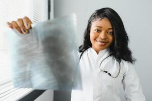 joven atractivo africano hembra médico radiólogo mirando a radiografía imagen, mientras en pie en ligero moderno oficina en clínica foto