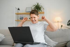 Happy excited grey haired mature woman celebrating online win, using laptop, looking at screen, sitting on couch at home, middle aged female feeling amazed, surprised by unbelievable good news. photo